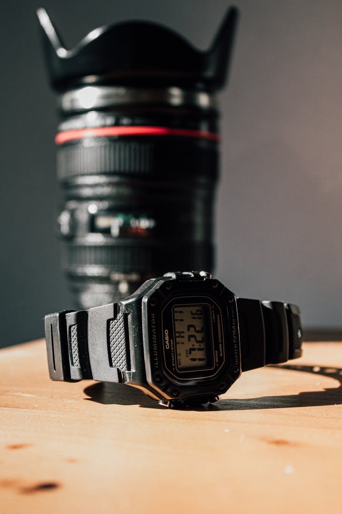 Digital wristwatch and camera lens on a wooden surface with sunlight.